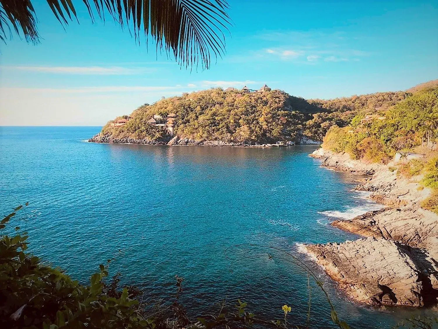Paisaje marino con agua turquesa, costa rocosa y una exuberante colina verde bajo un cielo azul brillante.