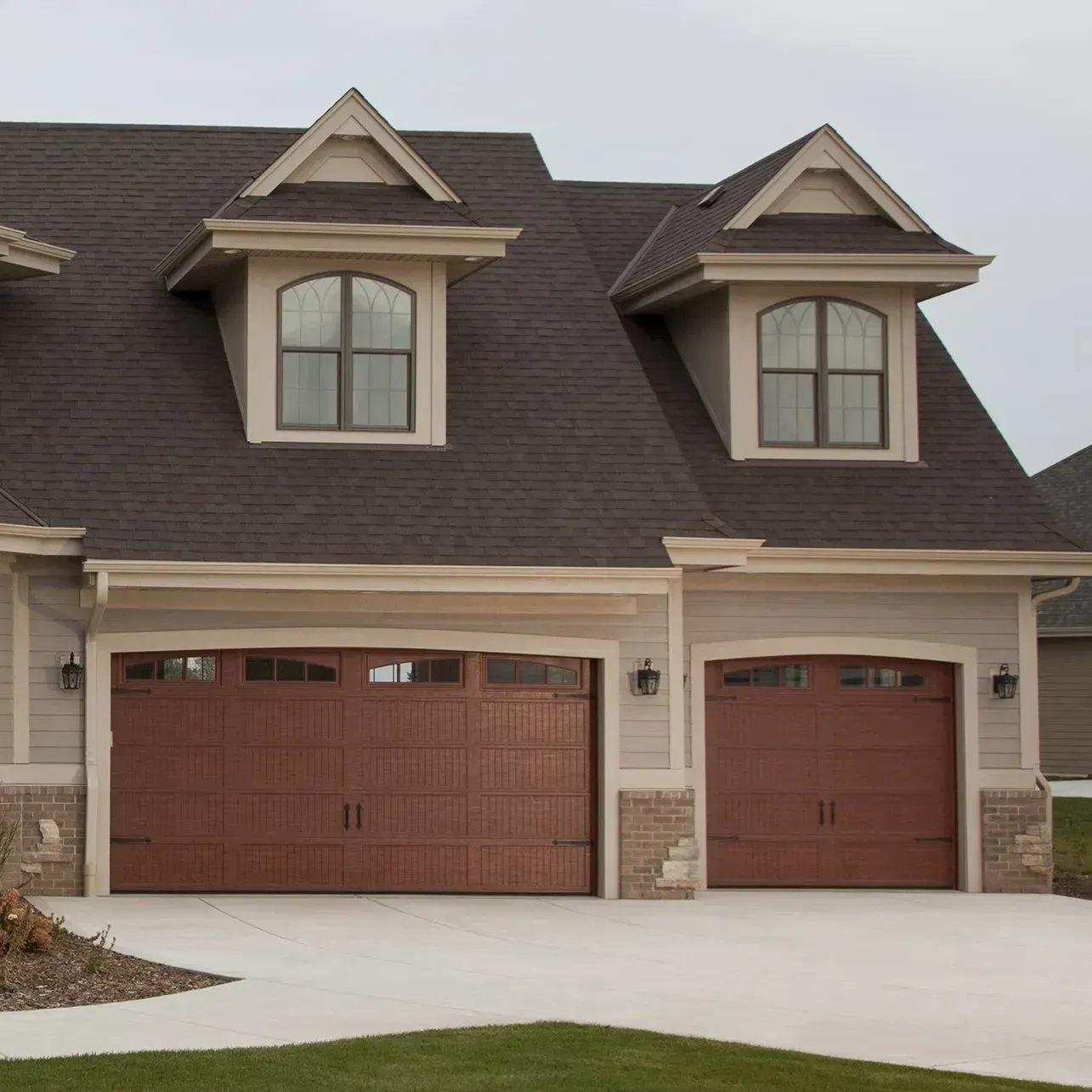 Brown garage doors with arched windows, light tan siding, and a dark brown roof.