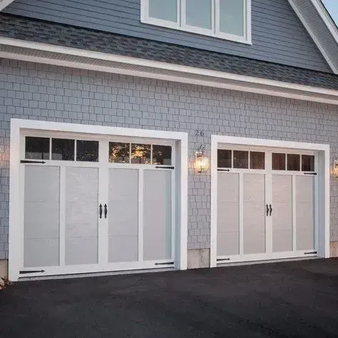 Two white garage doors with glass panels, set in a gray shingled house, asphalt driveway.