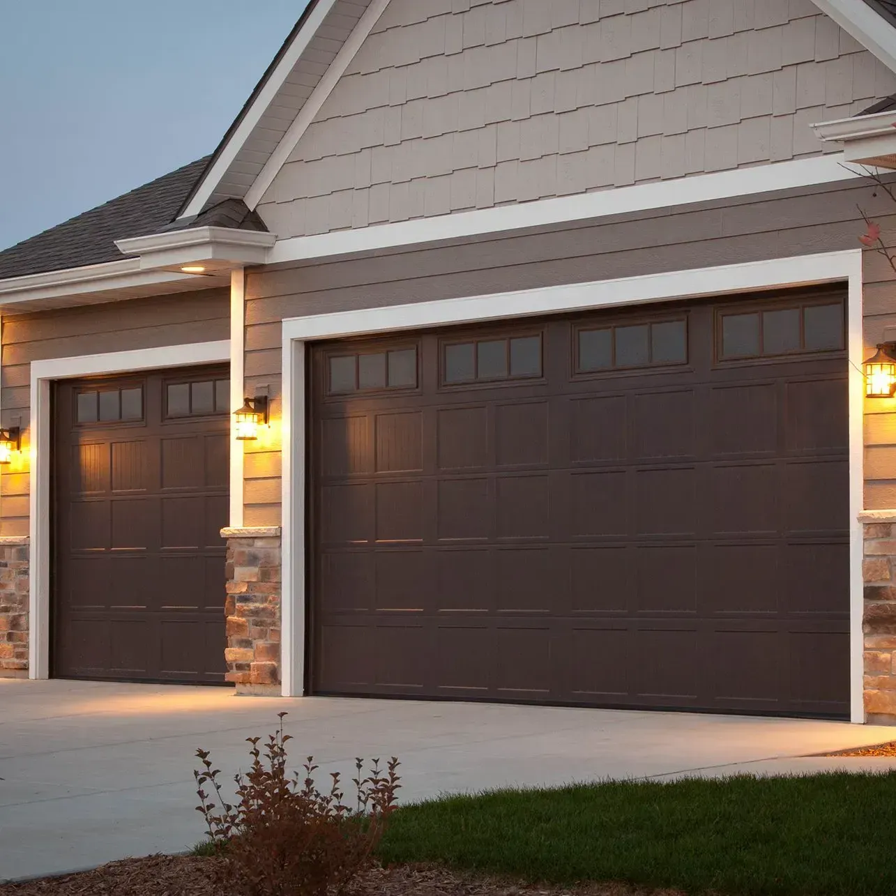 Brown garage doors with windows on a light beige house at dusk, lit by exterior lights.
