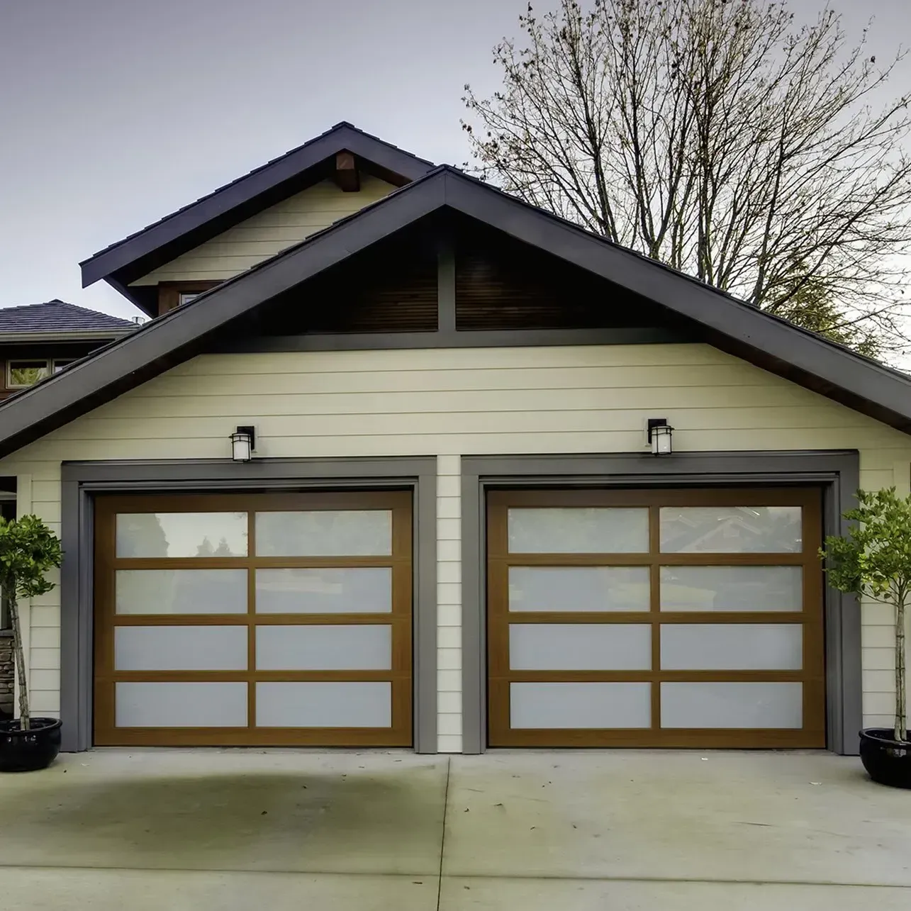 Two brown-framed garage doors with frosted glass panels on a beige house; small trees in pots flank each door.