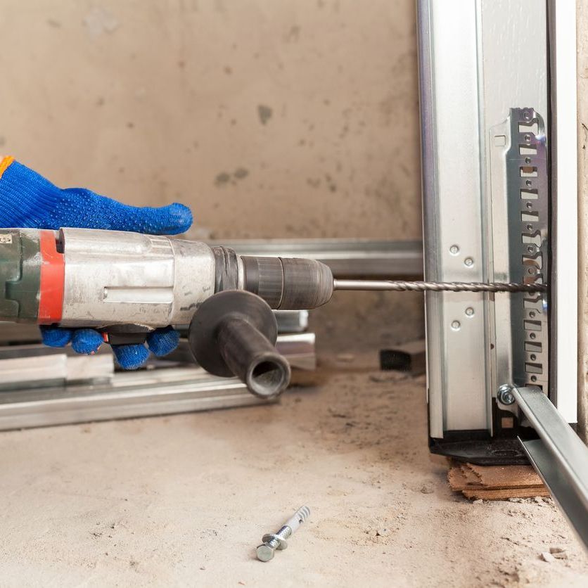 A gloved hand uses a drill on a metal garage door frame.