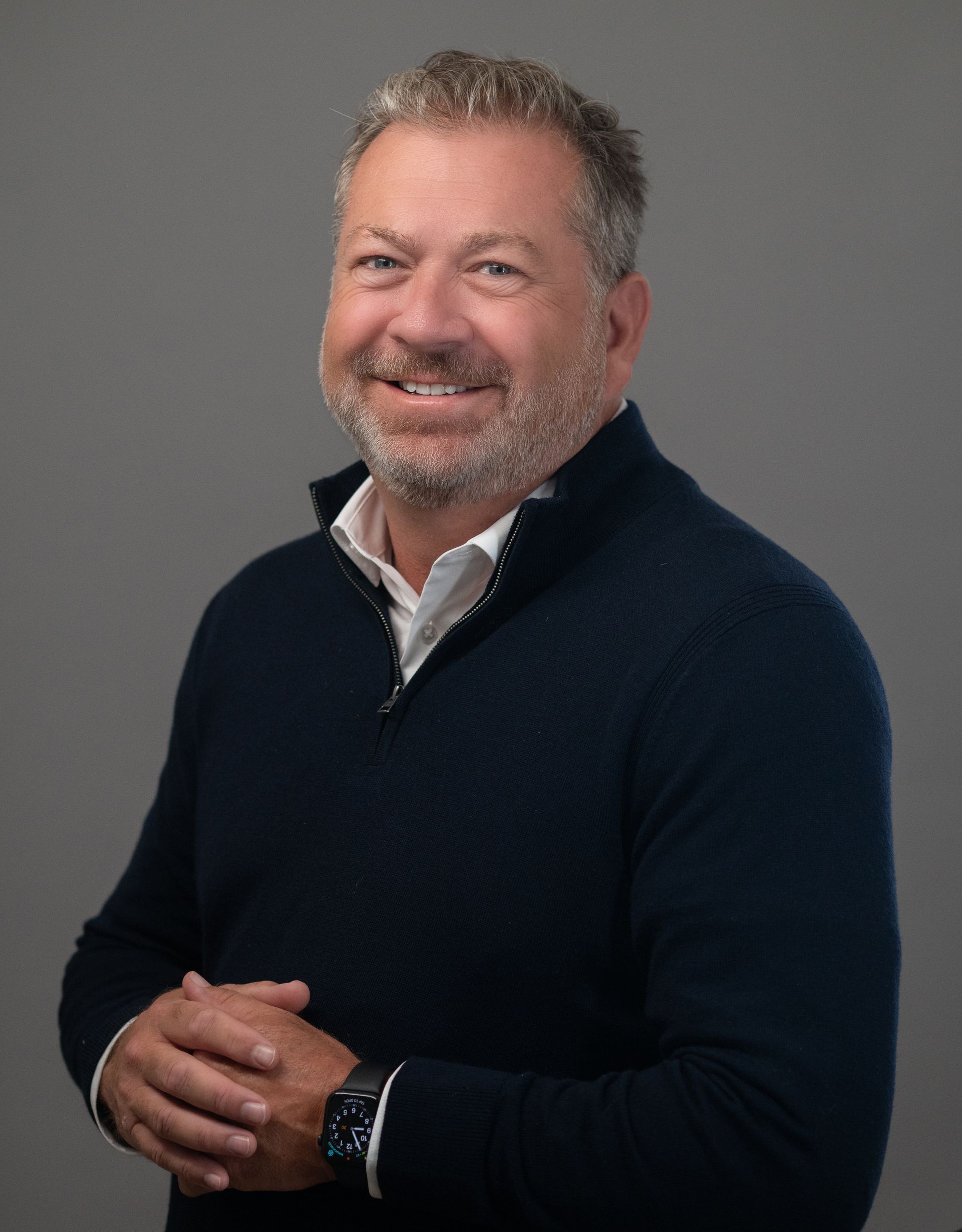 Man in navy sweater smiles, clasps hands, white shirt collar visible; gray background.