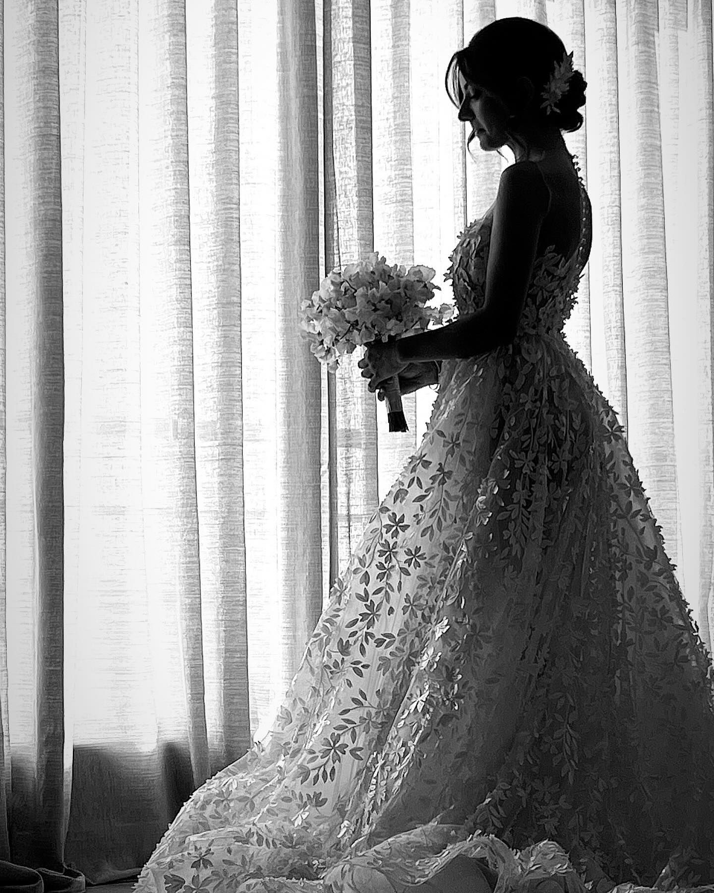 Una sposa in abito da sposa è in piedi davanti a una finestra con in mano un mazzo di fiori.
