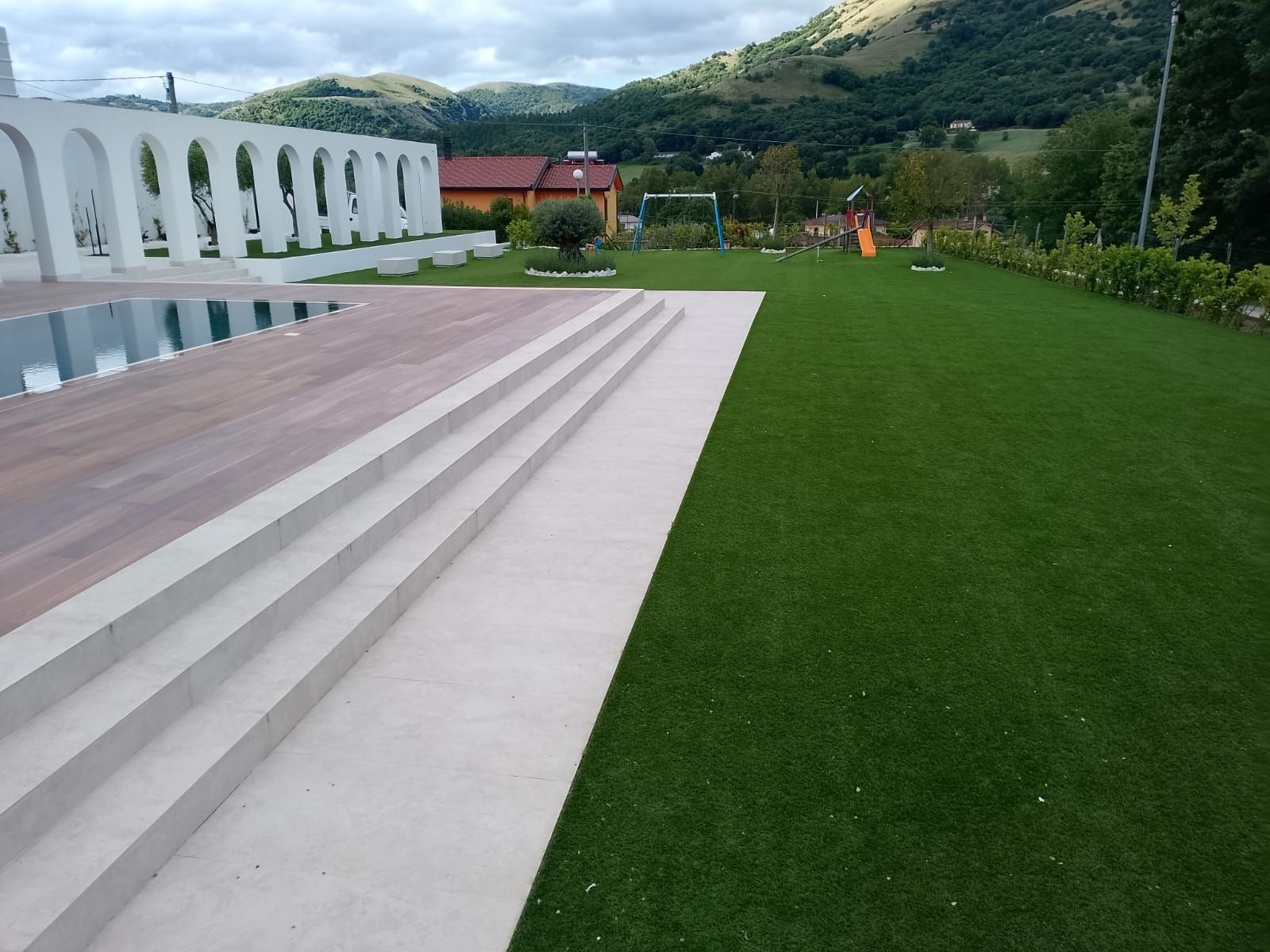 Una terrazza con gradini in pietra chiara conduce a una piattaforma in legno accanto a una piscina, delimitata da un prato verde ben curato.