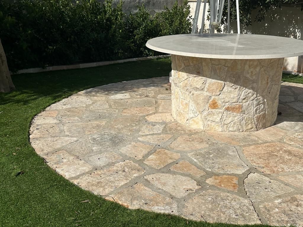 Un patio circolare pavimentato in pietra presenta un tavolo cilindrico rialzato in pietra, immerso in un giardino con prato verde.