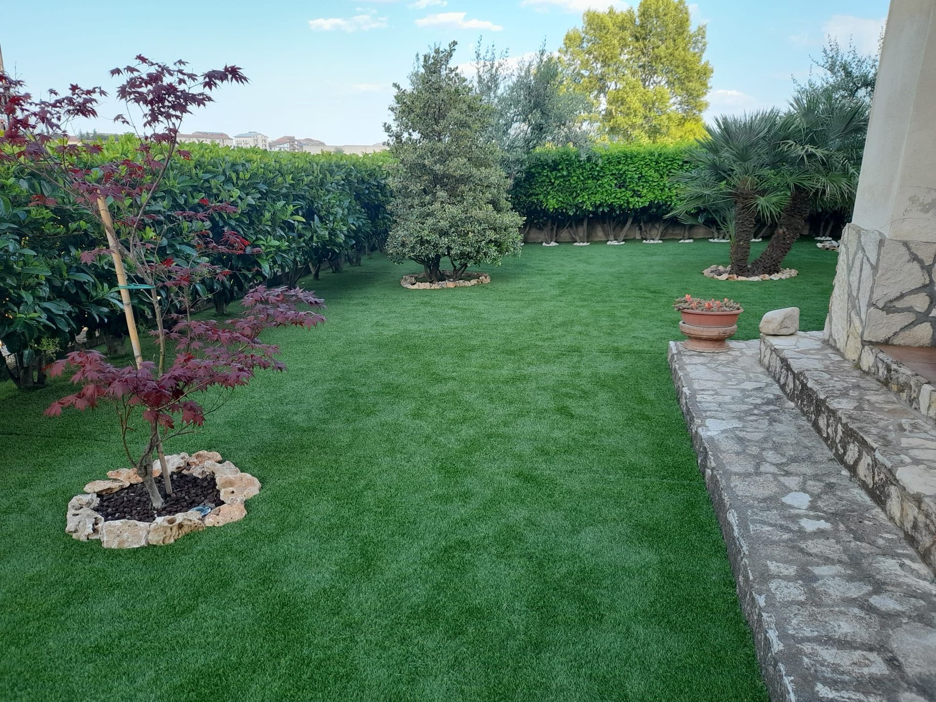 Un prato verde brillante con piccoli alberi, siepi e un vialetto lastricato in pietra accanto a un edificio con muri in pietra.