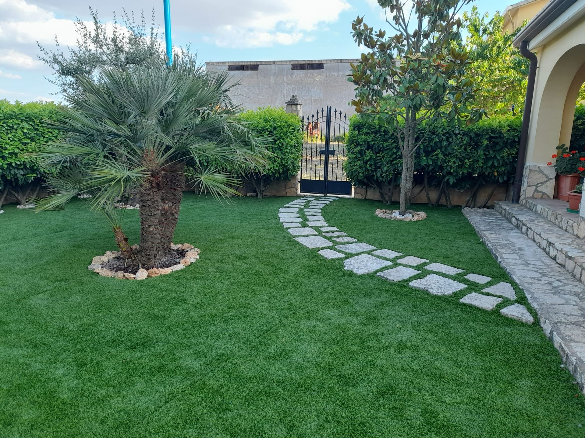 Un rigoglioso prato verde con una piccola palma, un sentiero di pietra, siepi ben curate e l'ingresso ad arco di una casa in pietra.