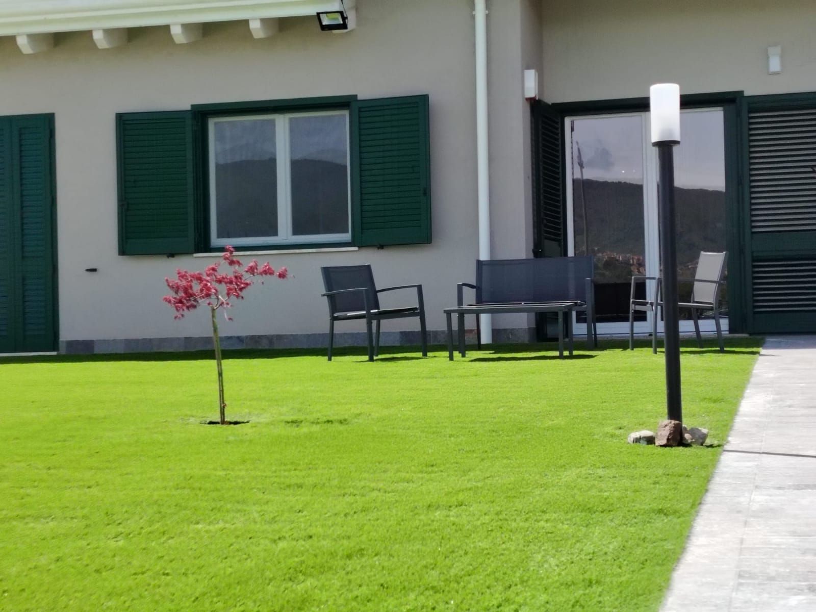 Area patio esterna con prato verde, sedie scure, un piccolo albero e un lampione di fronte a una casa con persiane verdi.