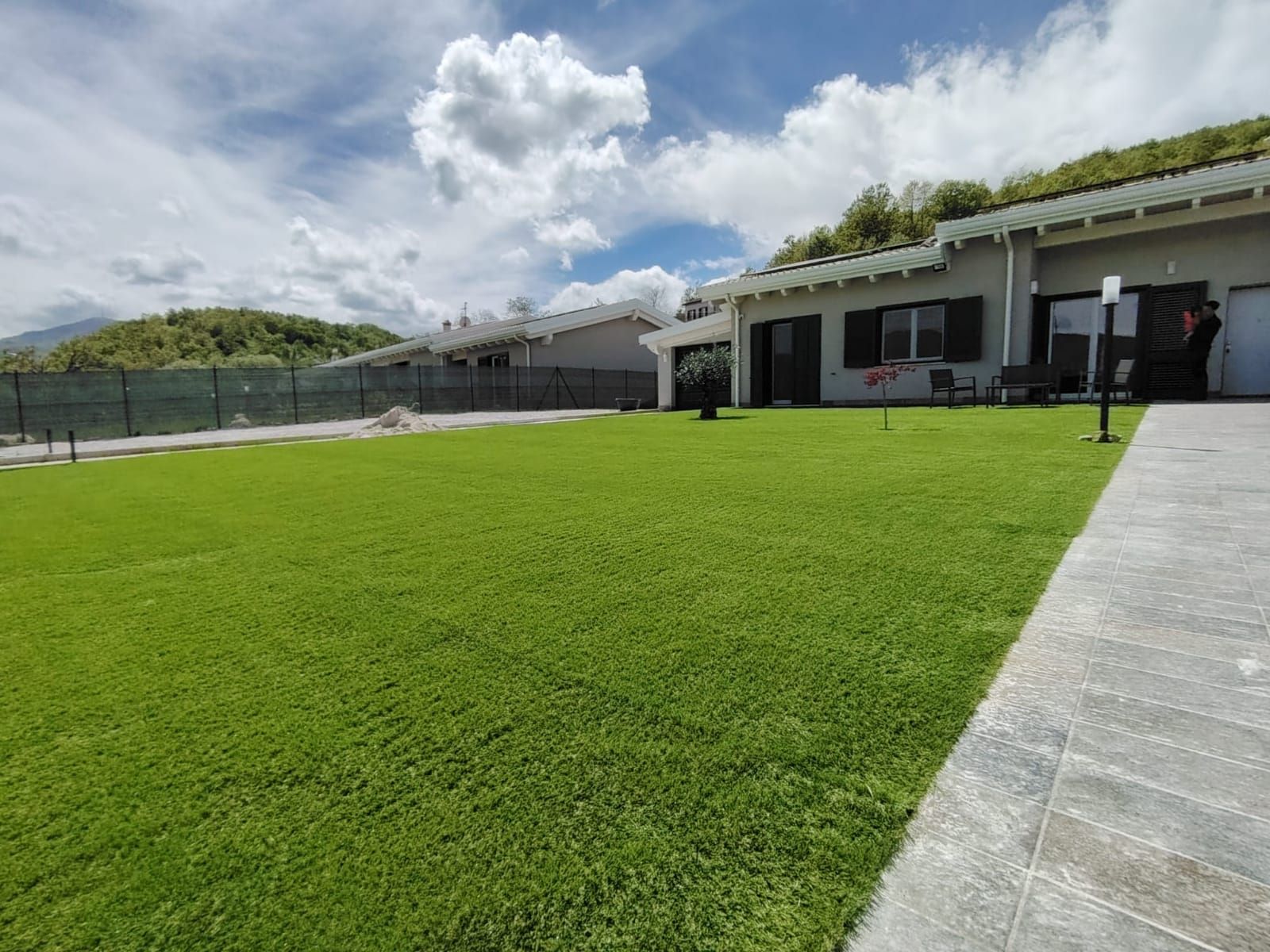 Un prato verde ben curato di fronte a una moderna casa a un piano con patio in pietra, sotto un cielo luminoso e nuvoloso.