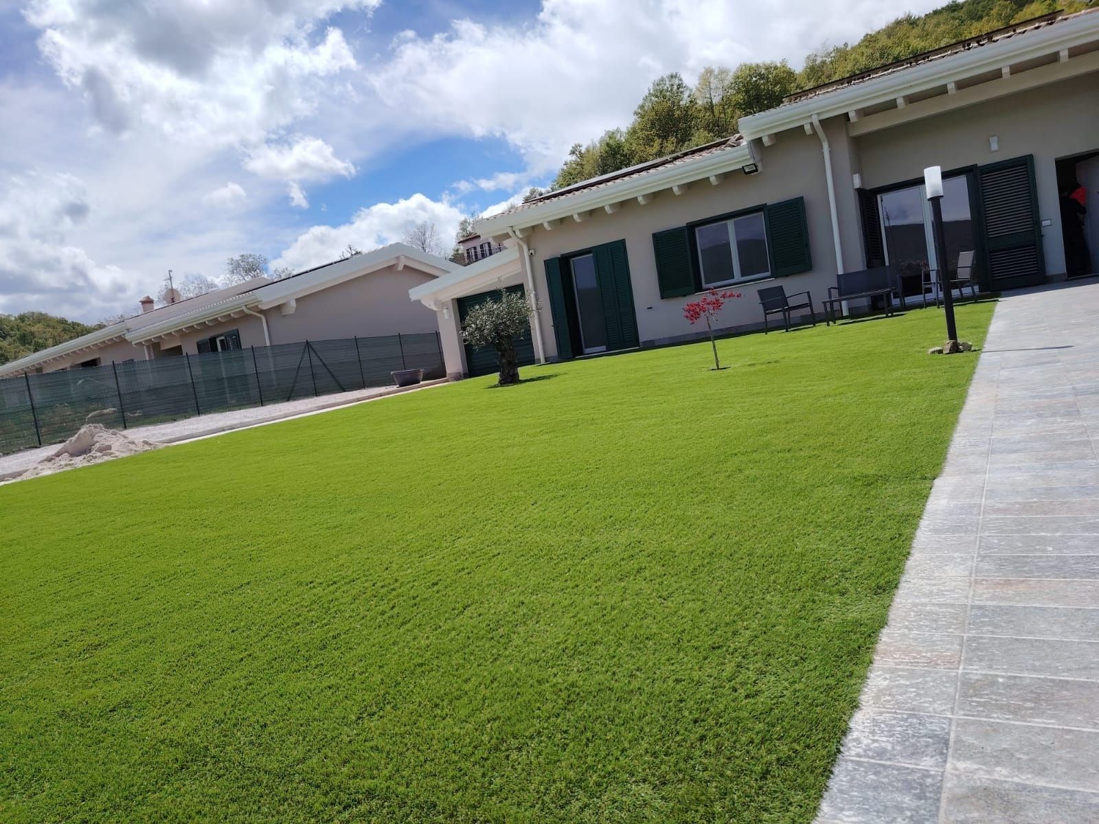 Un prato verde brillante conduce a un edificio basso con persiane verde scuro e a un vialetto di pietra sotto un cielo parzialmente nuvoloso.