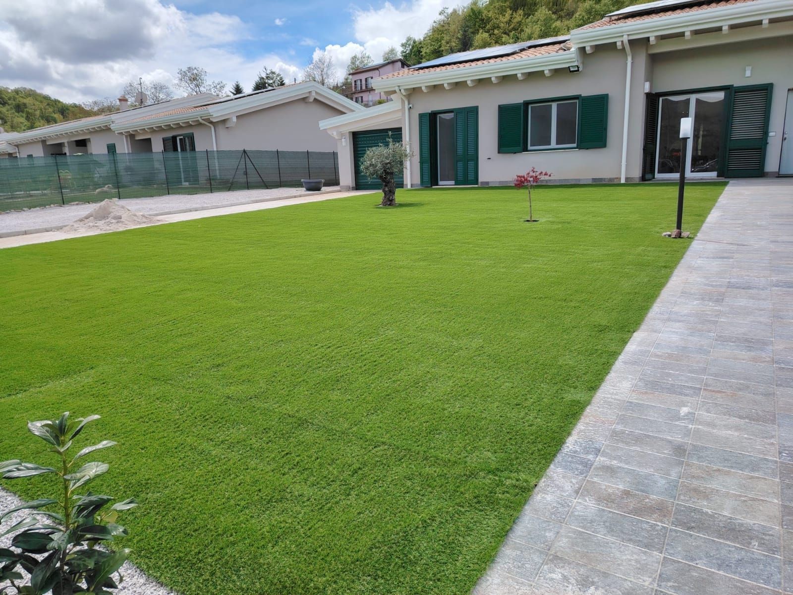 Un rigoglioso prato verde si estende verso una casa a un solo piano con persiane verdi e un vialetto di pietra sotto un cielo azzurro nuvoloso.