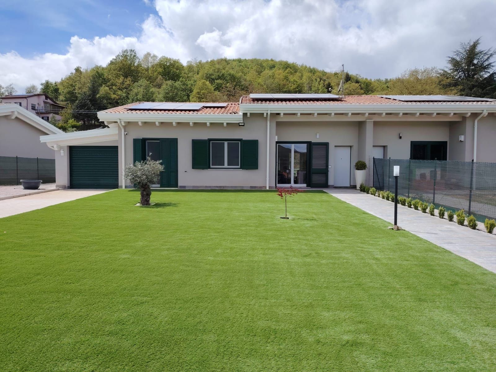 Una casa a un solo piano, di colore chiaro, con un prato verde, persiane scure, pannelli solari e un vialetto lastricato sotto un cielo azzurro.