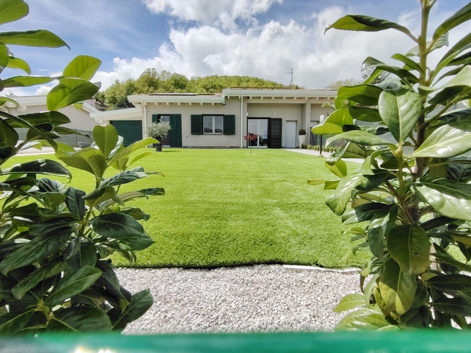 Una casa beige a un piano con persiane verdi, sullo sfondo di un prato verde ben curato e incorniciata da rigogliosi cespugli verdi.