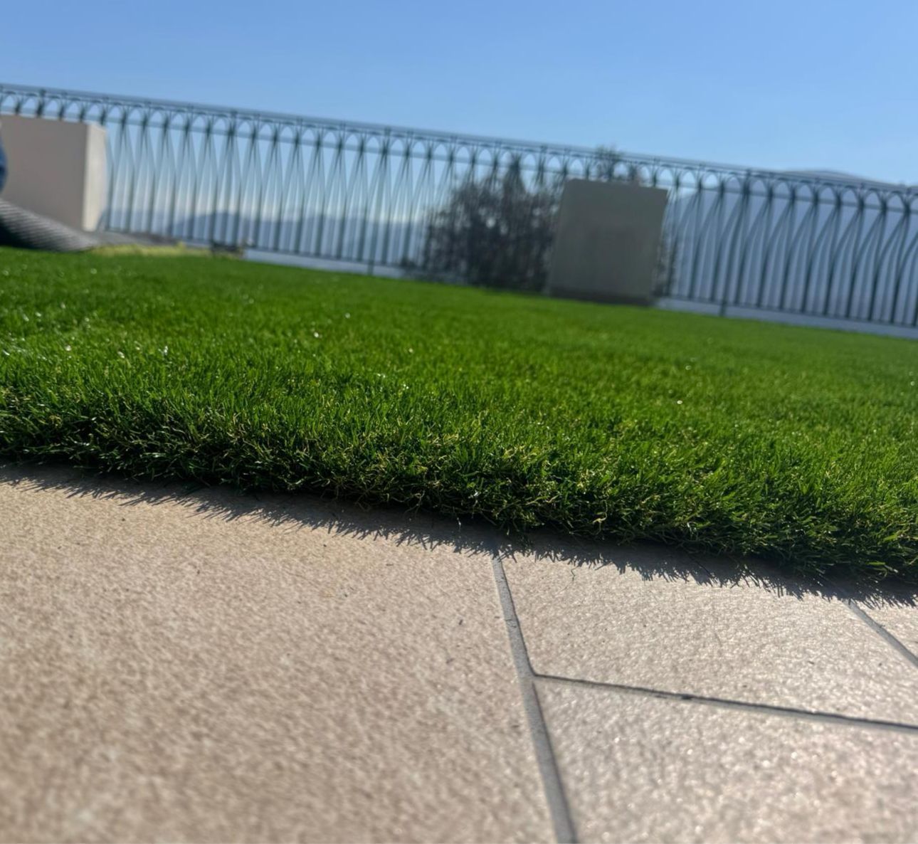 Una porzione di erba sintetica verde che sfuma in piastrelle di pietra chiara, con una ringhiera metallica e un cielo limpido sullo sfondo.