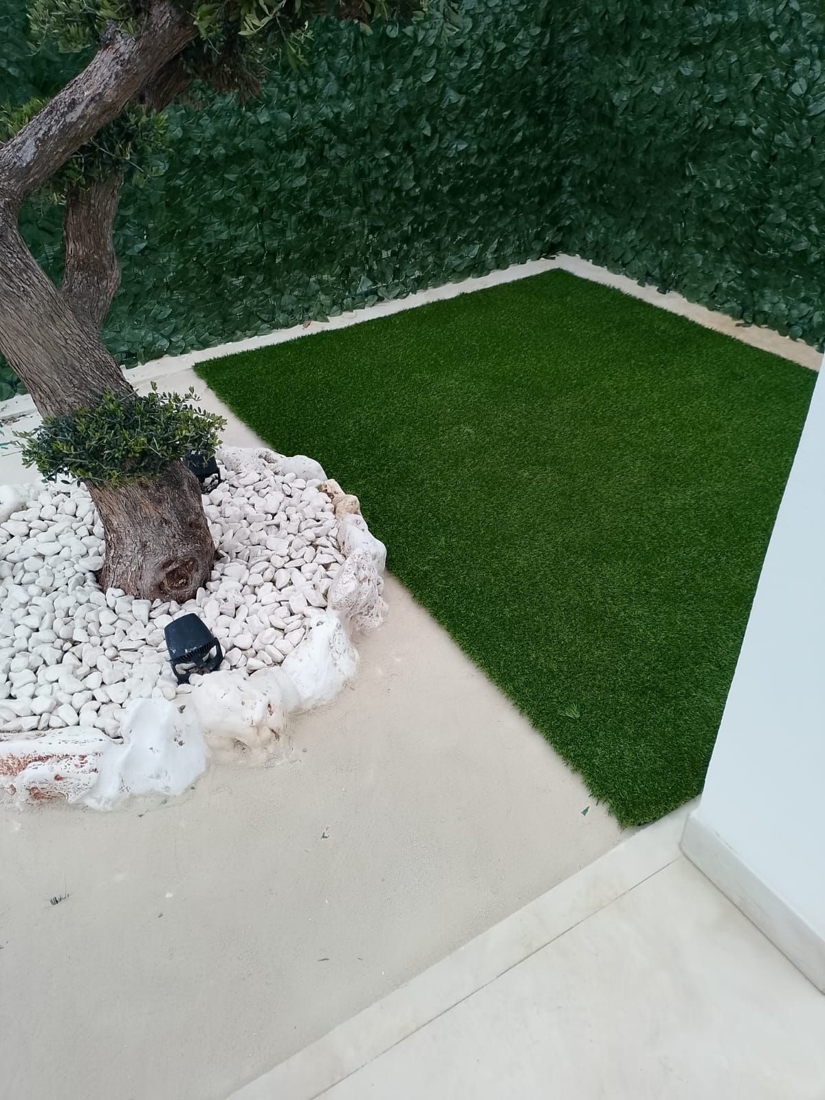 Un patio con un albero circondato da pietre bianche, adiacente a una zona quadrata di erba sintetica e a una siepe verde che garantisce la privacy.