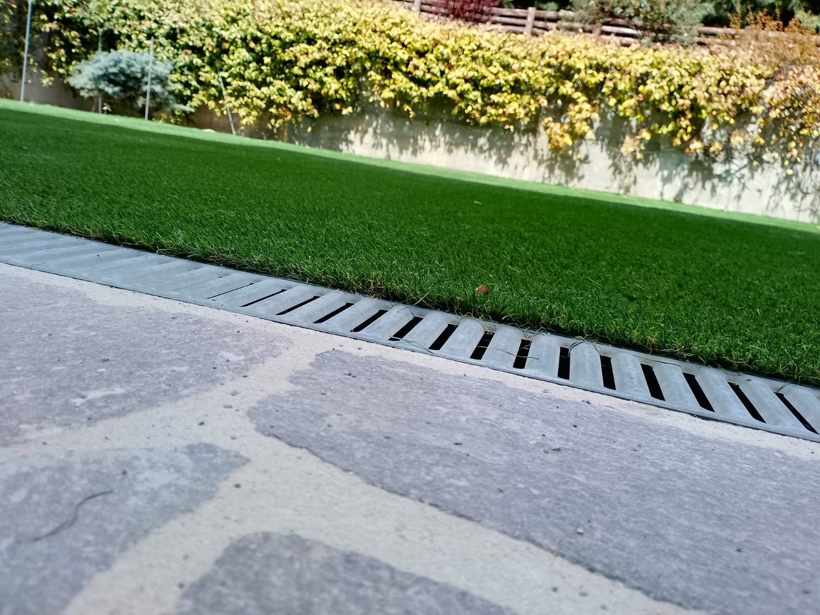 Un patio in pietra separa una griglia di drenaggio pavimentata da una zona di erba sintetica verde, con una siepe sullo sfondo.