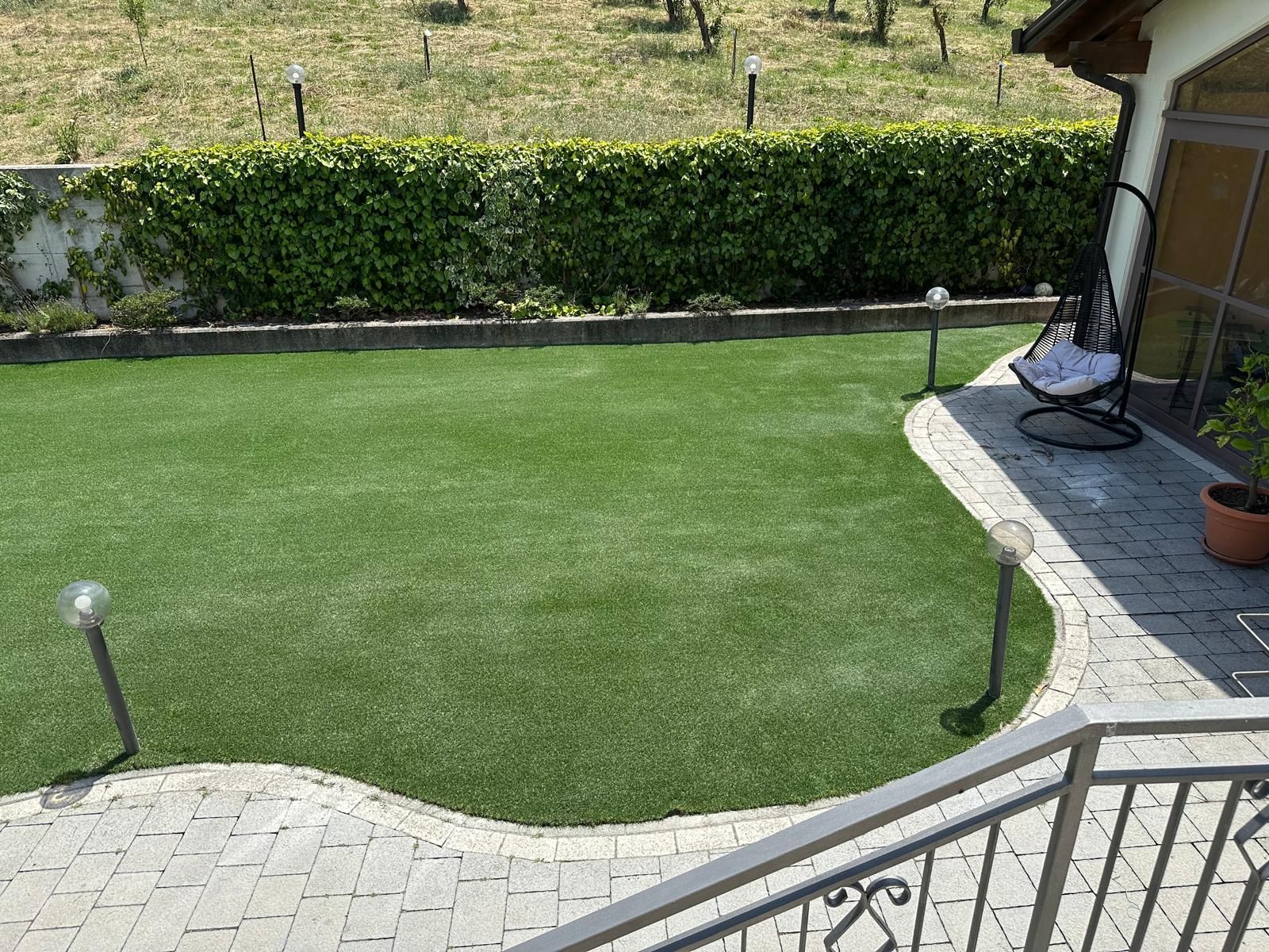 Un prato verde ben curato, incorniciato da lastre di pietra grigia, un'alta siepe e luci esterne, con una sedia sospesa sul patio.