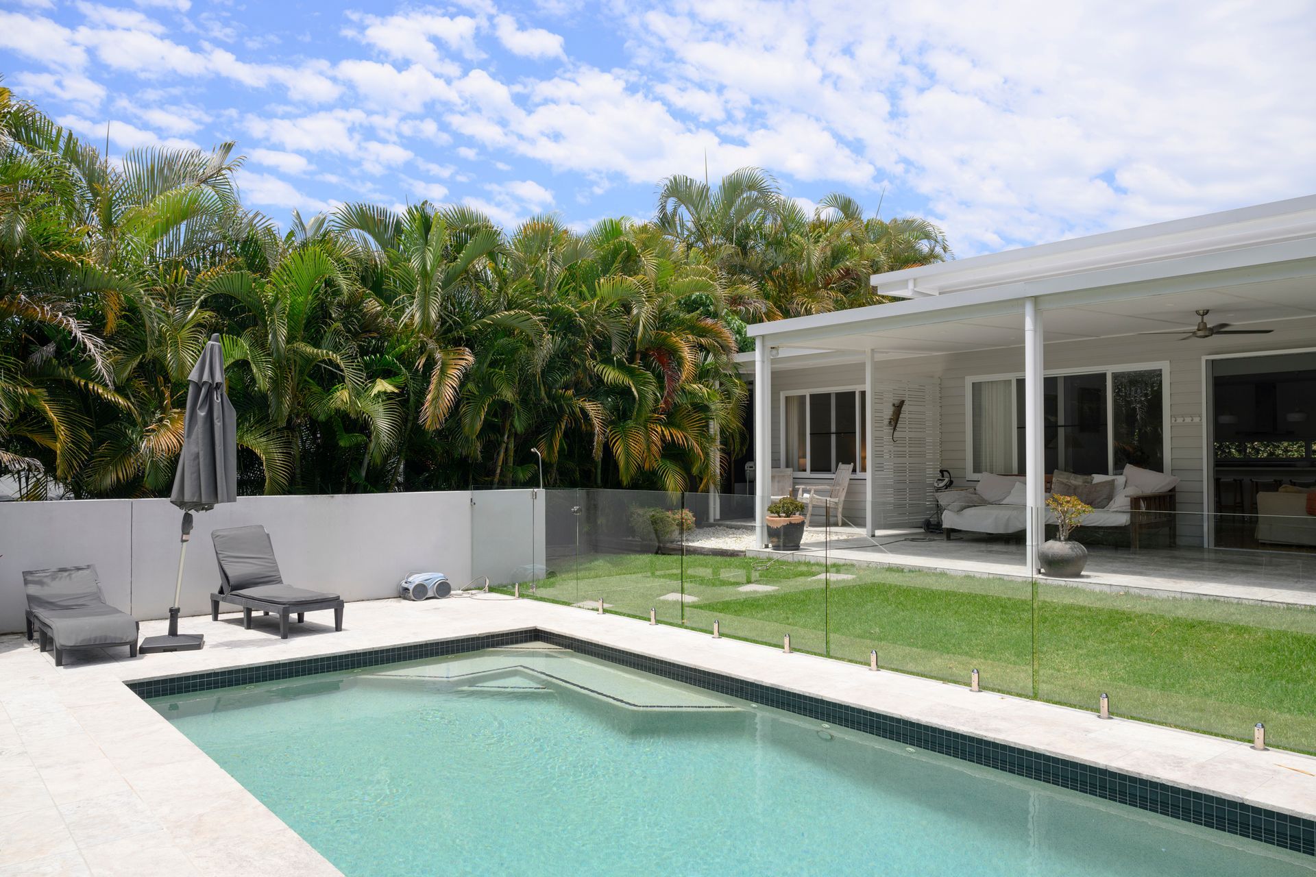 Home with enclosed patio, lawn, poolside lounge chairs and umbrella, and lush palm grove.