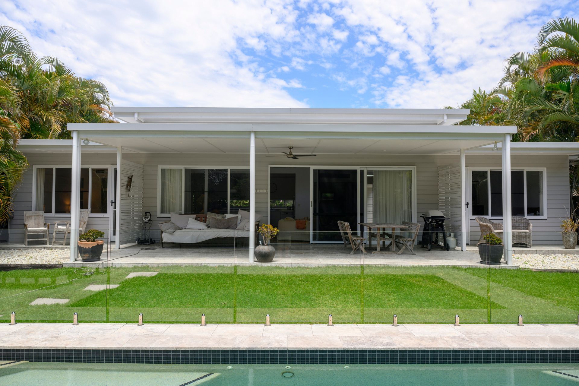 View across swimming pool enclosed in glass, green lawn, covered patio, and outdoor furniture.