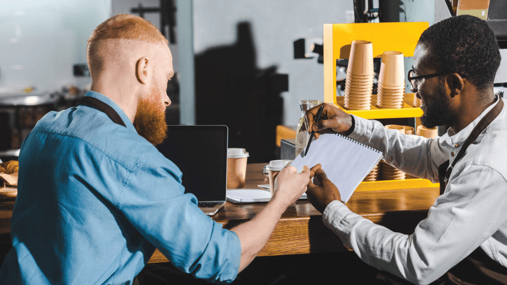 Two men are sitting at a table in a cafe looking at a piece of paper.