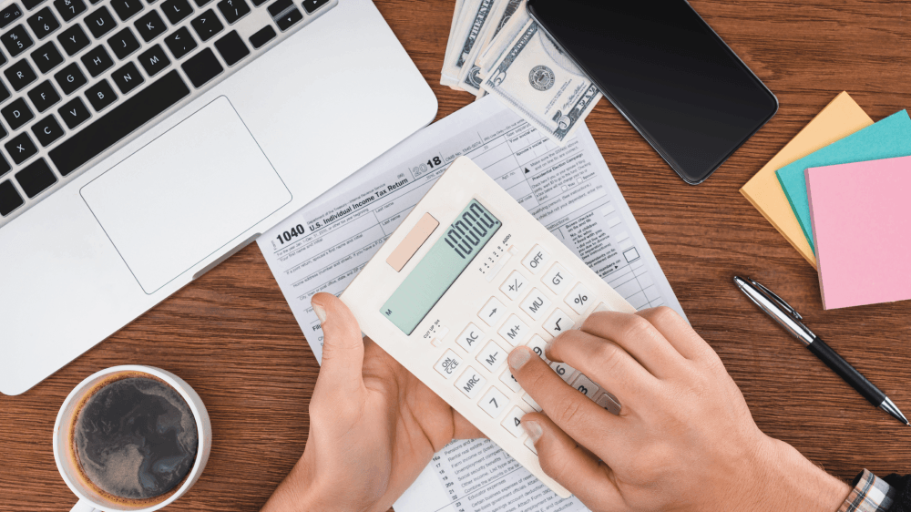 A person is holding a calculator in front of a laptop and a cup of coffee.