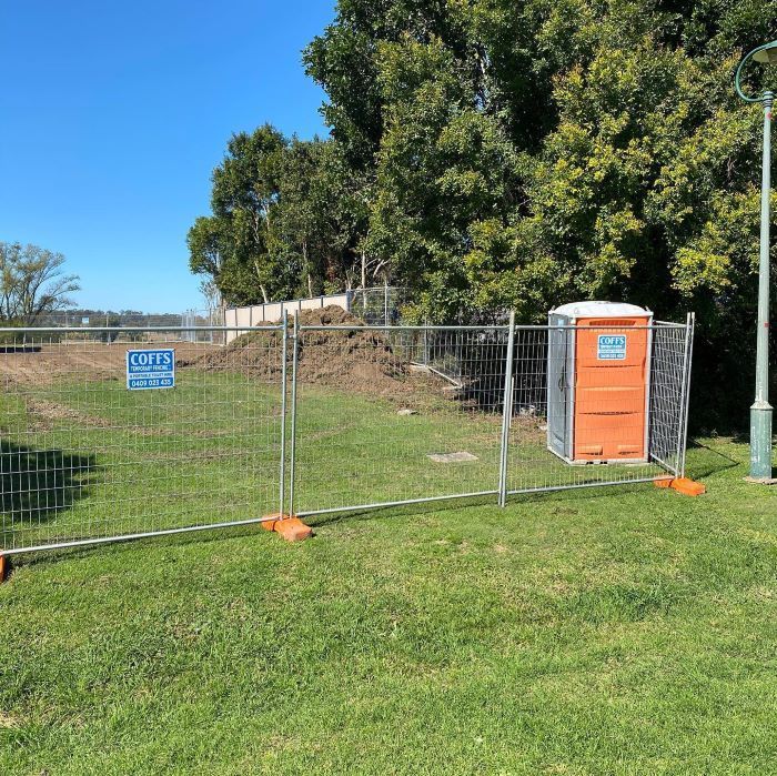 A Portable Toilet Is Behind A Fence In A Grassy Field — Coffs Temporary Fencing Pty Ltd In Moonee Beach, NSW