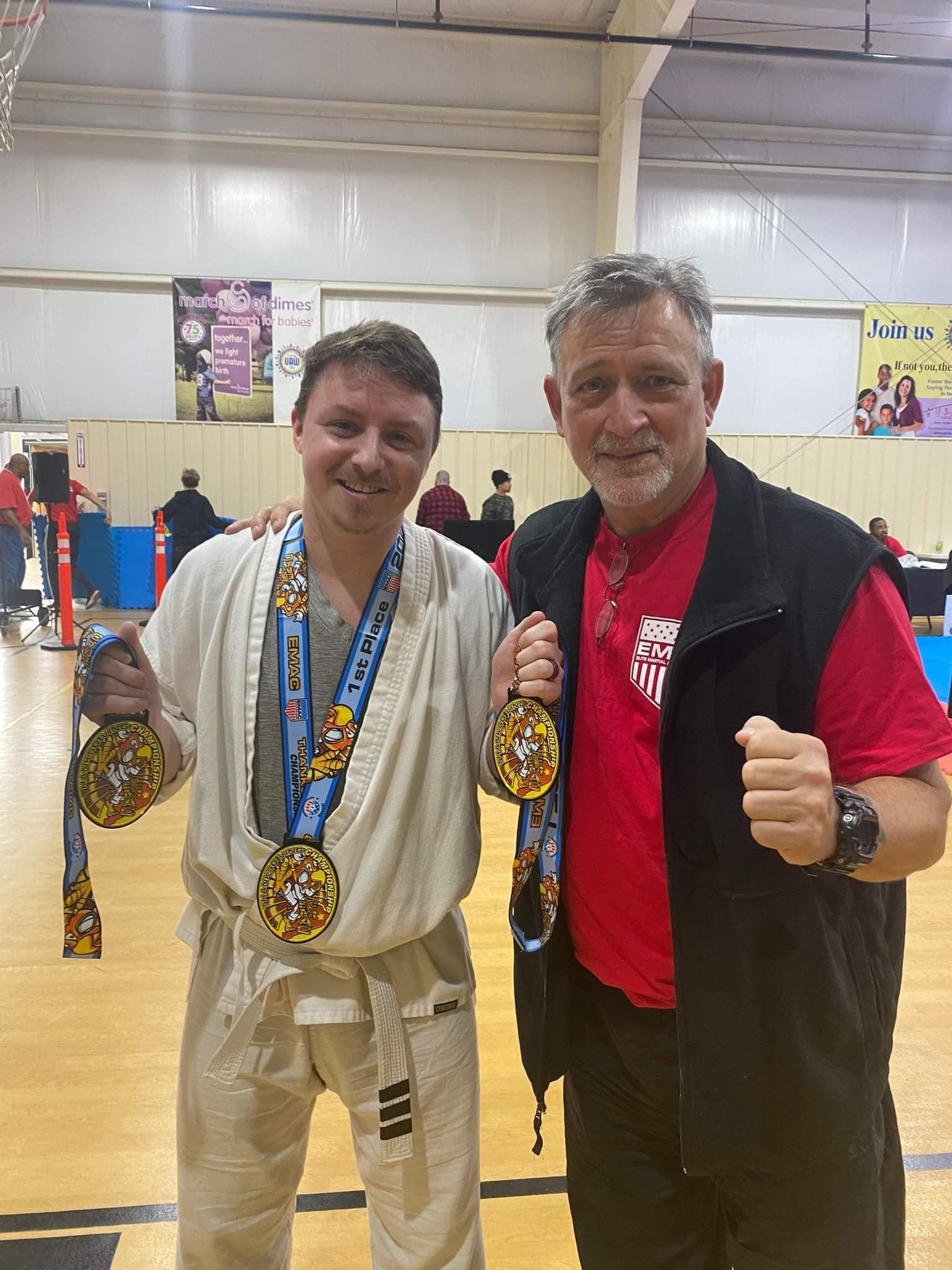 Two men are standing next to each other in a gym holding medals.