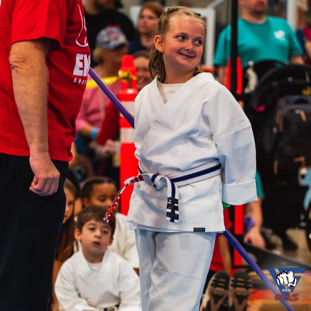 A little girl in a white karate uniform is smiling while standing next to a man in a red shirt that says en