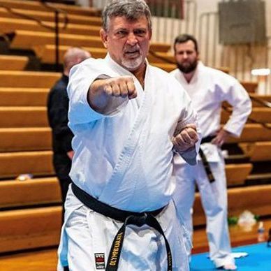 A man in a white karate uniform is standing on a blue mat in a gym.