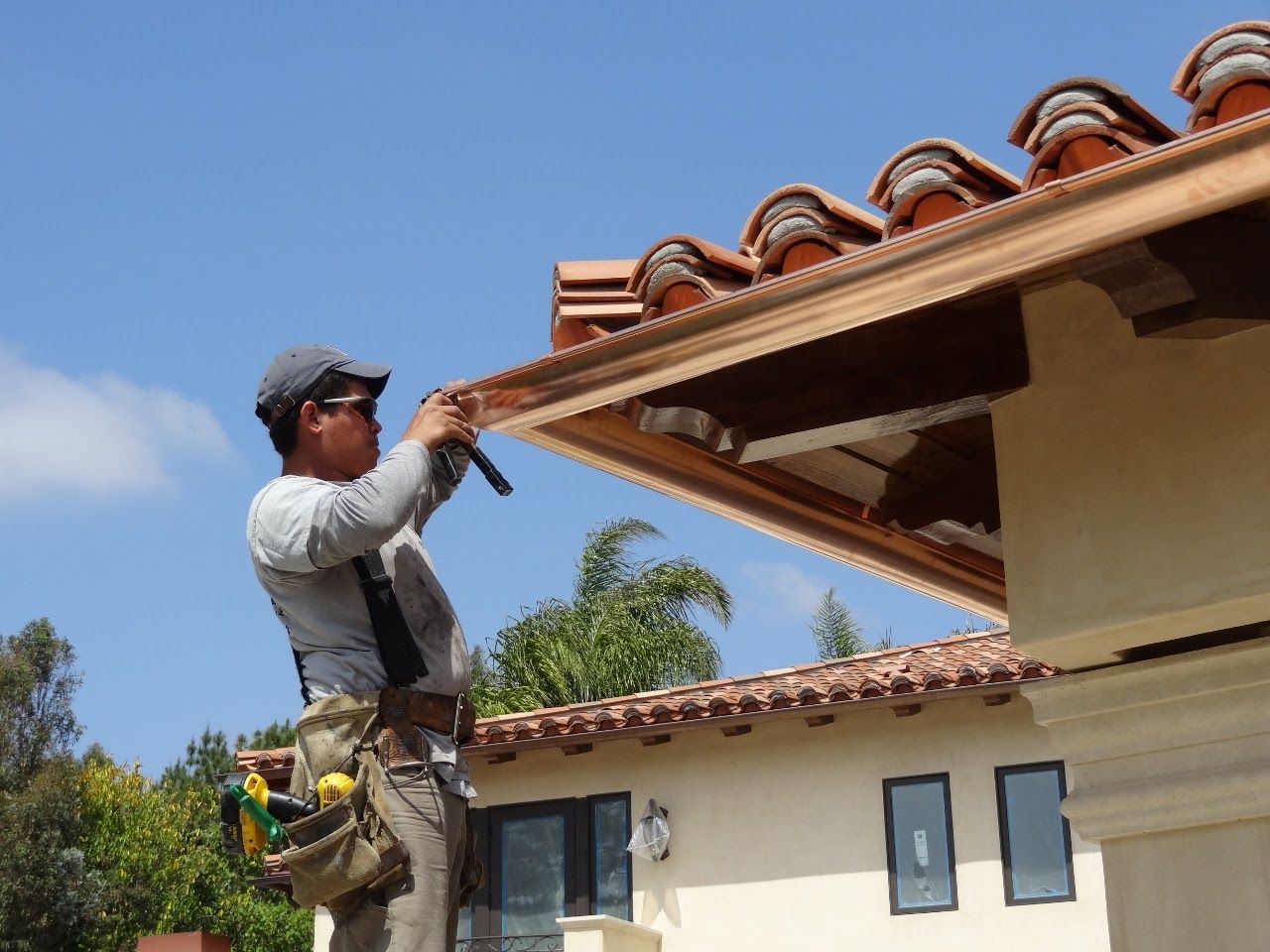 Man on a ladder installing copper gutters on a house with a Spanish-style tile roof.