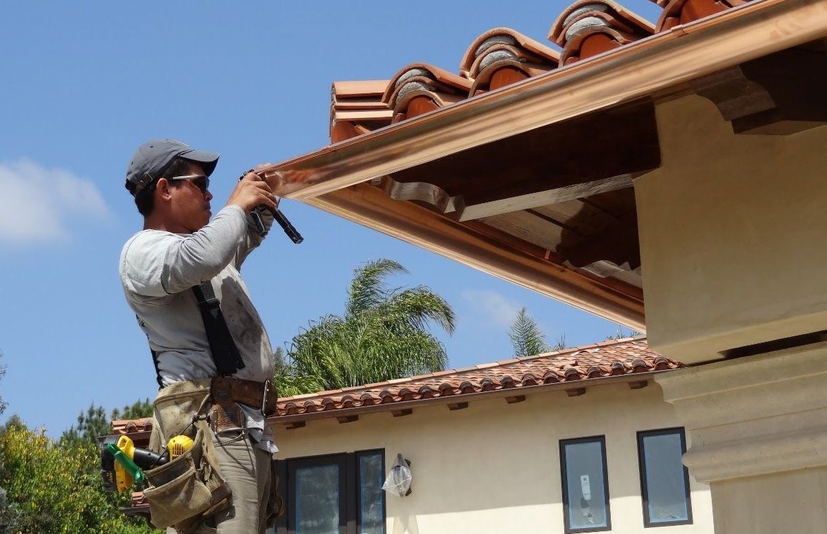 A man in safety gear installs gutters on a house with a terracotta roof on a sunny day.