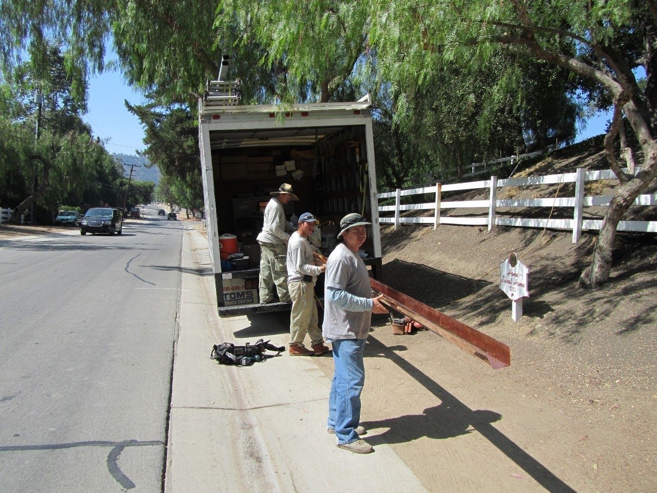 Men unloading wood from a truck on a sunny street near a white fence.