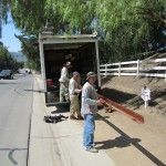 Three men unloading lumber from a truck on a roadside, next to a white fence and greenery.