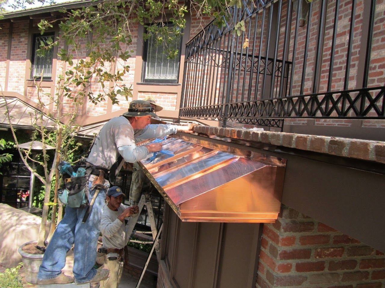 Workers install a copper gutter on a brick building with a balcony.