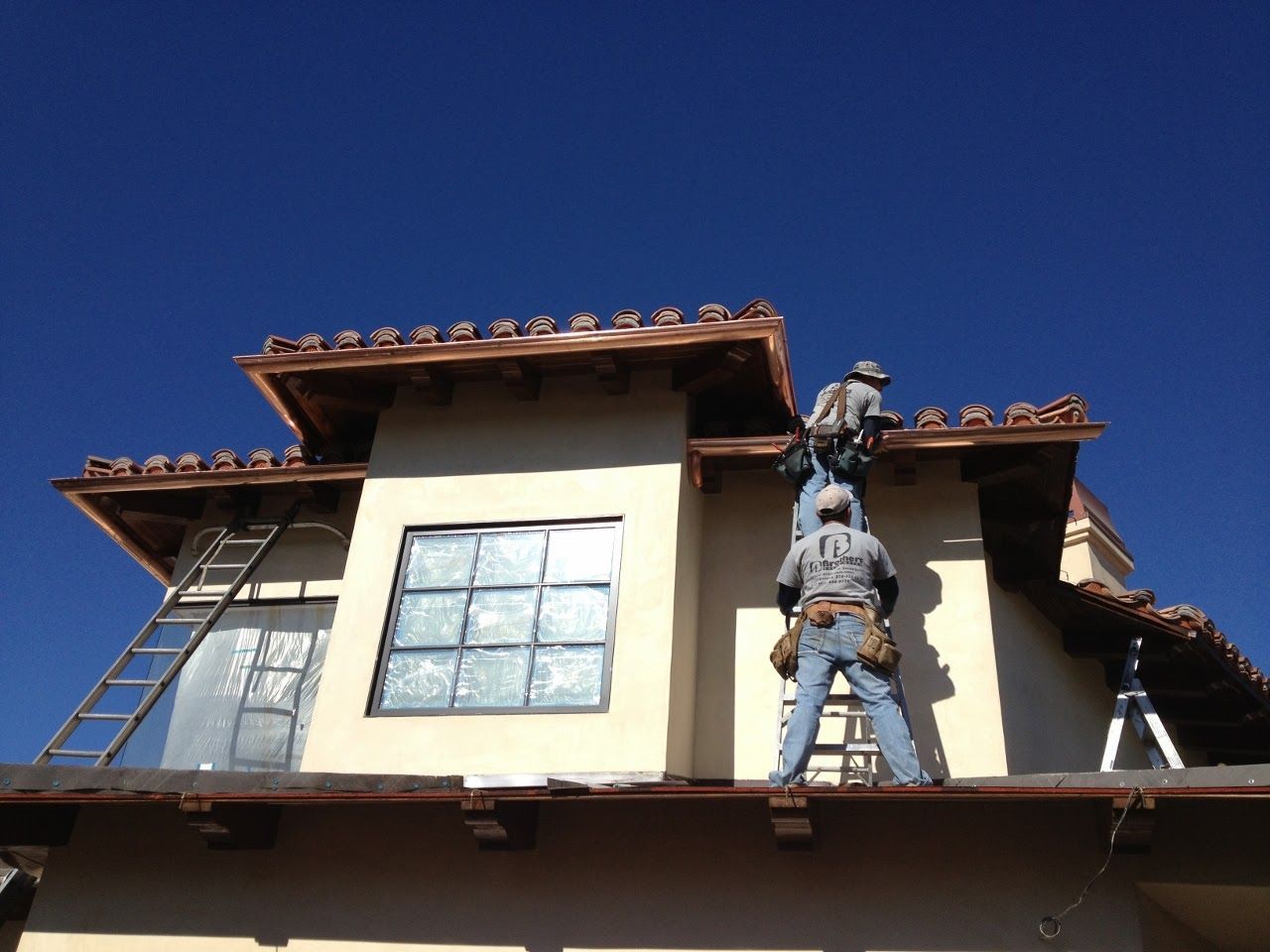 Two roofers installing copper gutters on a two-story house against a blue sky.