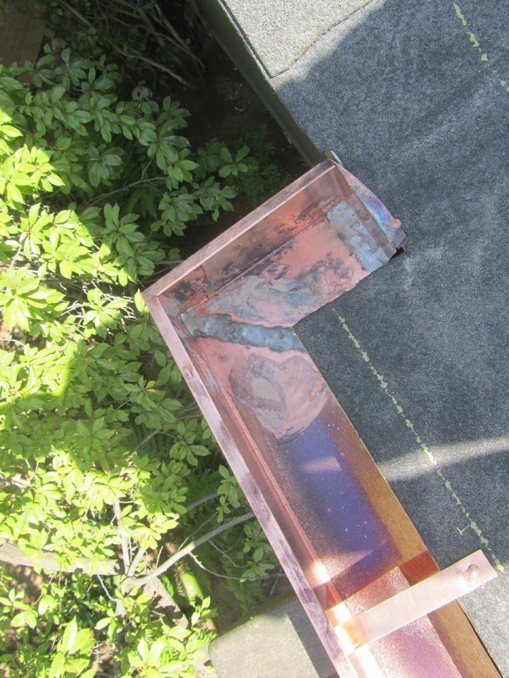 Copper gutter corner on a rooftop with faded reddish-brown metal and greenery in the background.