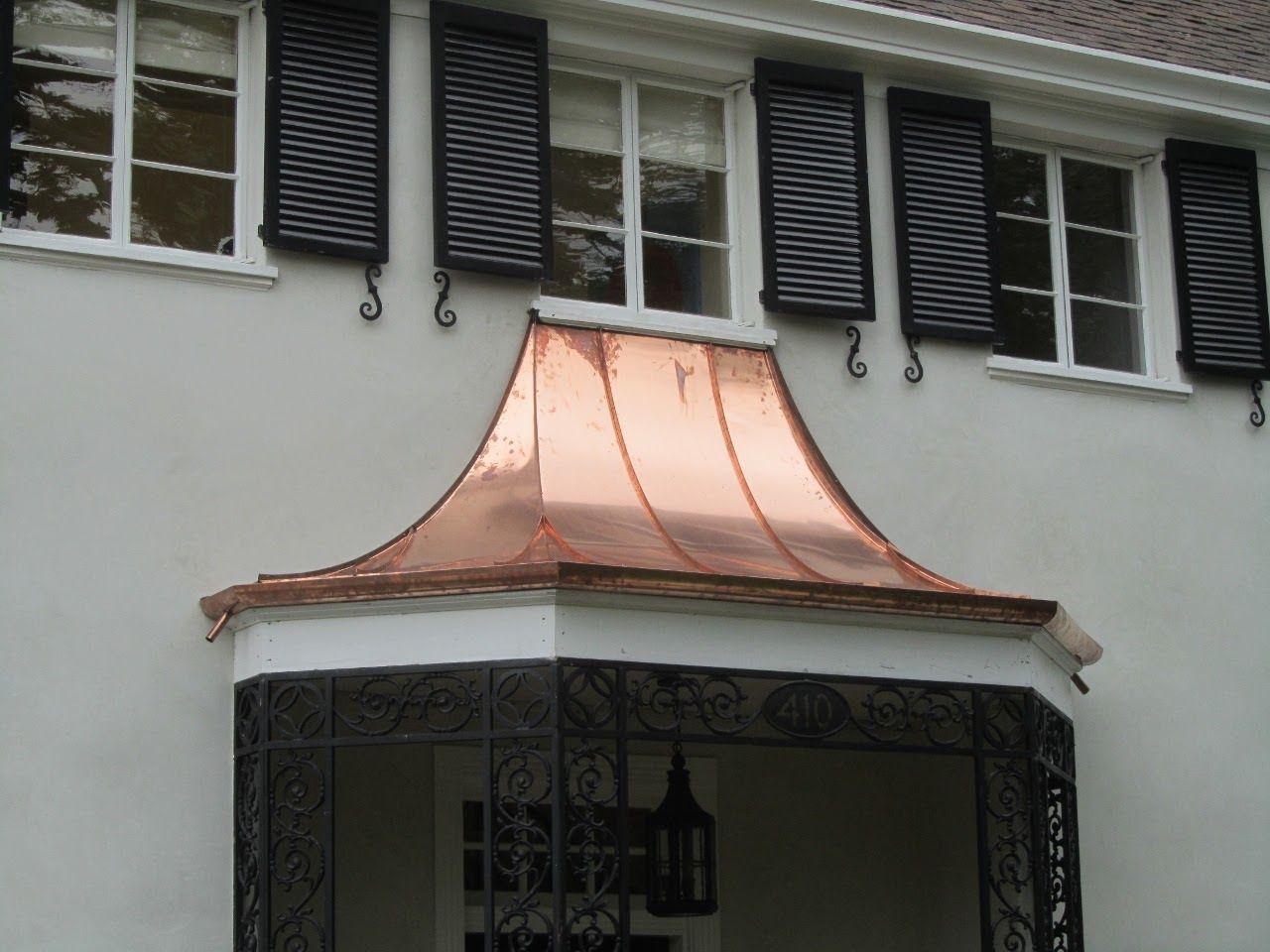 Copper roof over a small porch with ornate black ironwork. Light-colored stucco wall, black shutters.