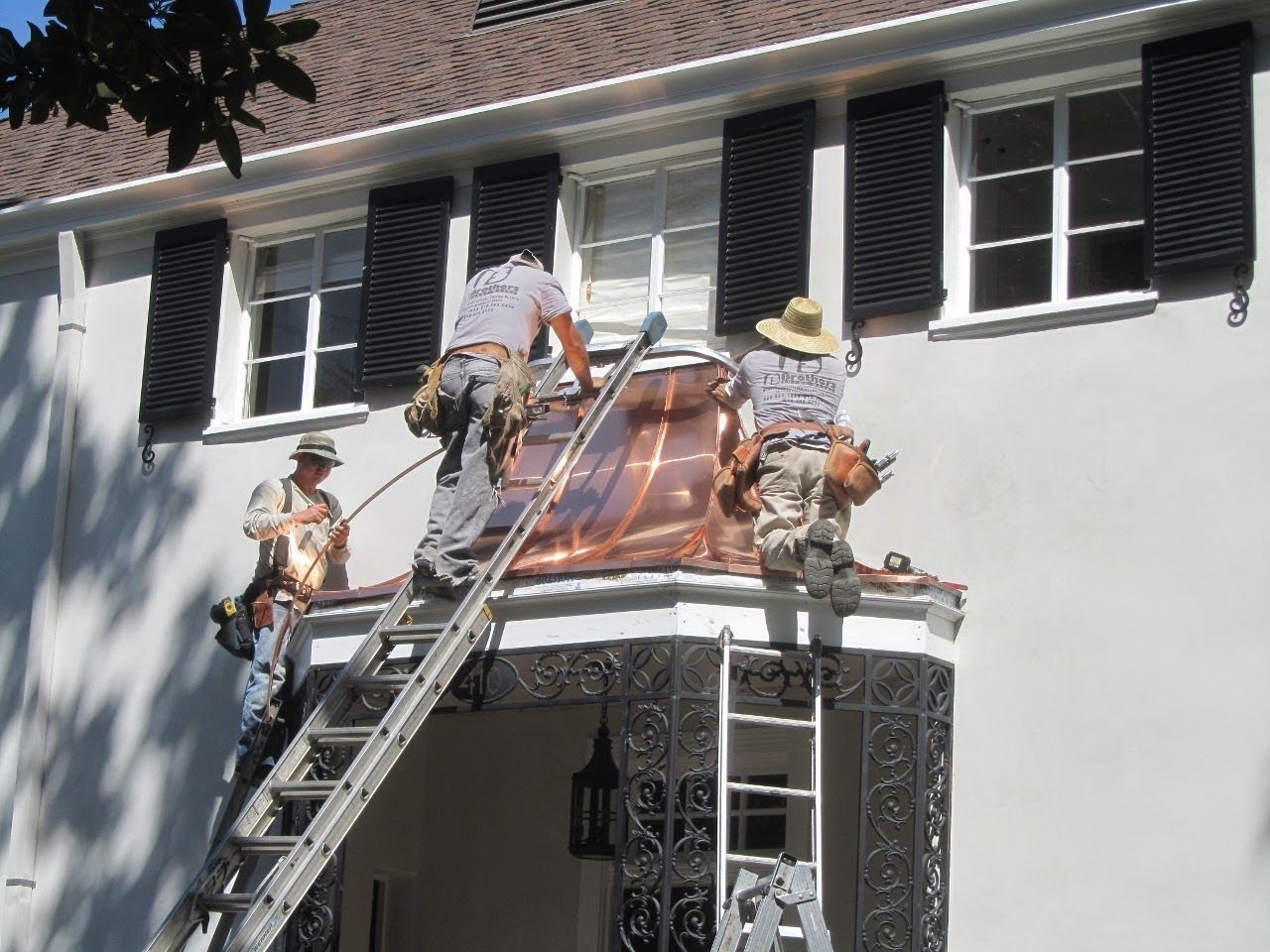 Construction workers installing copper roofing on a white building with black shutters.