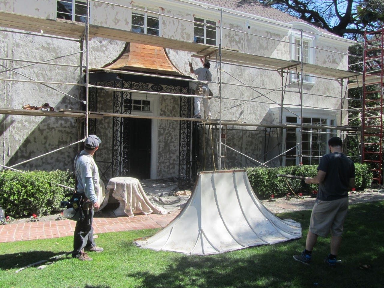 Exterior house under renovation with scaffolding, workers, and large fabric canopy on grass.
