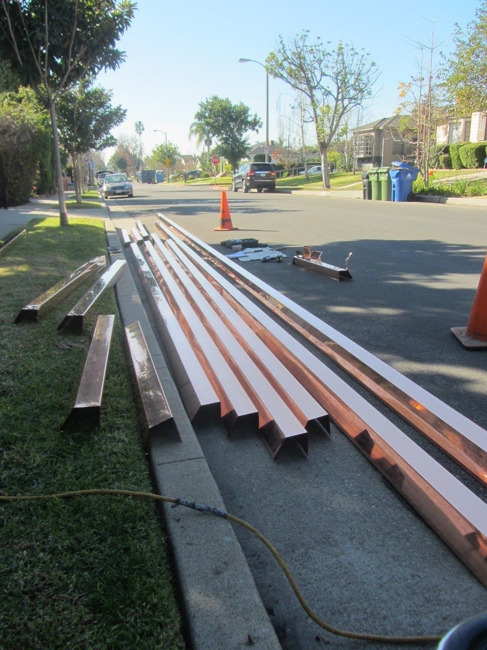 Copper gutter materials laid on a sidewalk next to a street with houses and a blue sky.