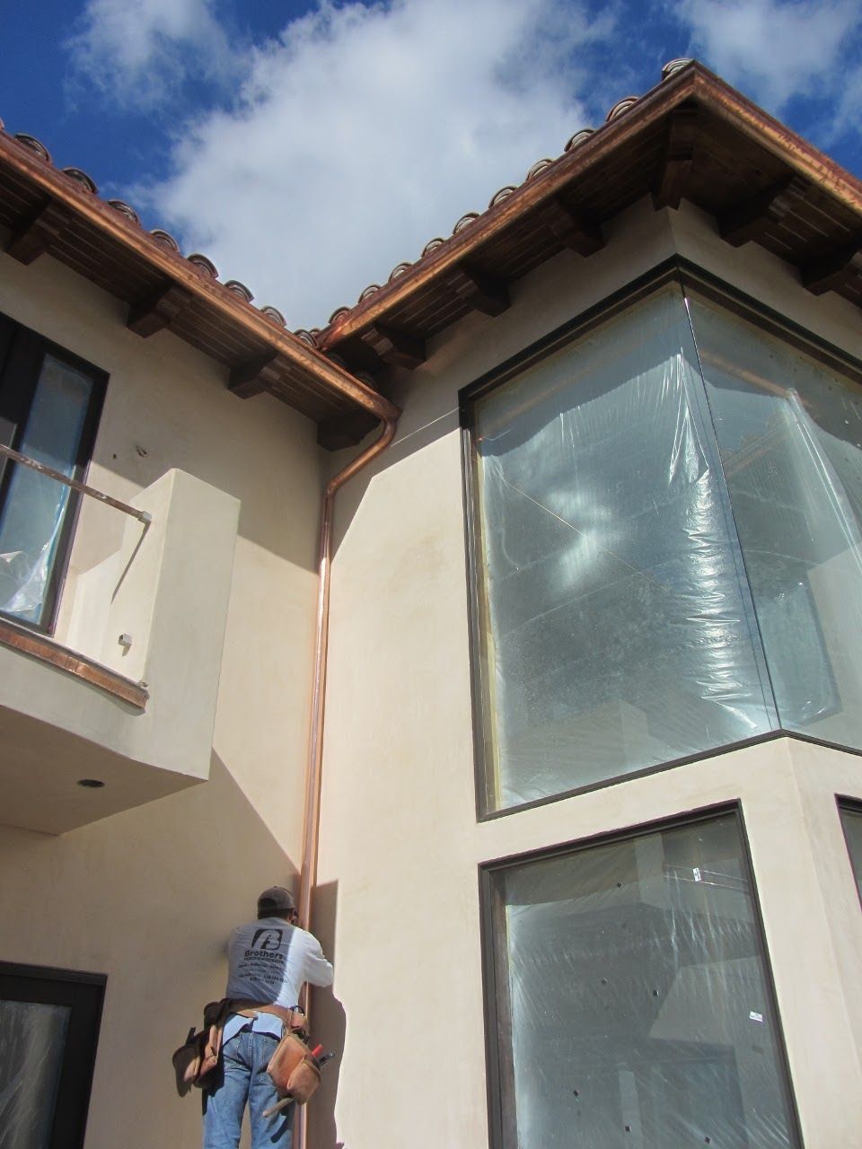 Man on a ladder installing copper gutters on a light-colored building with large windows and a tiled roof.