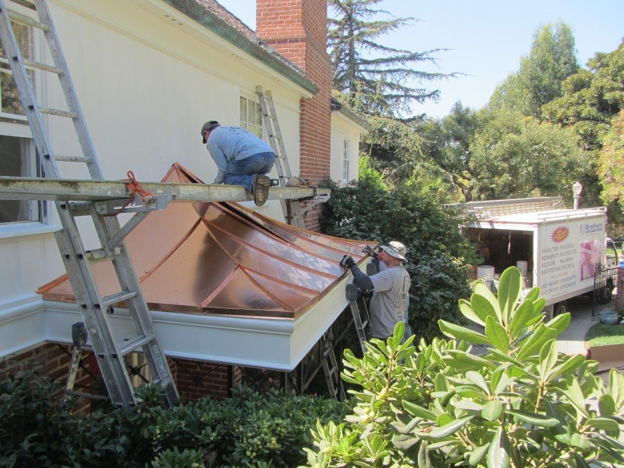 Two workers install copper roof on house bump-out; white stucco wall, brick base, ladders, truck, trees.