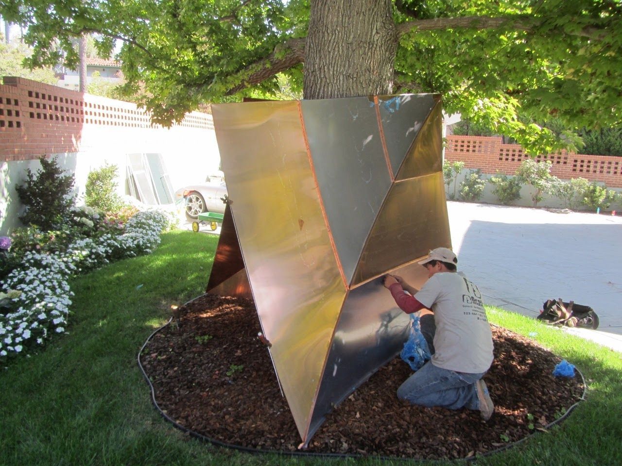 Man installing copper geometric art around a tree, outdoors, in a grassy yard.