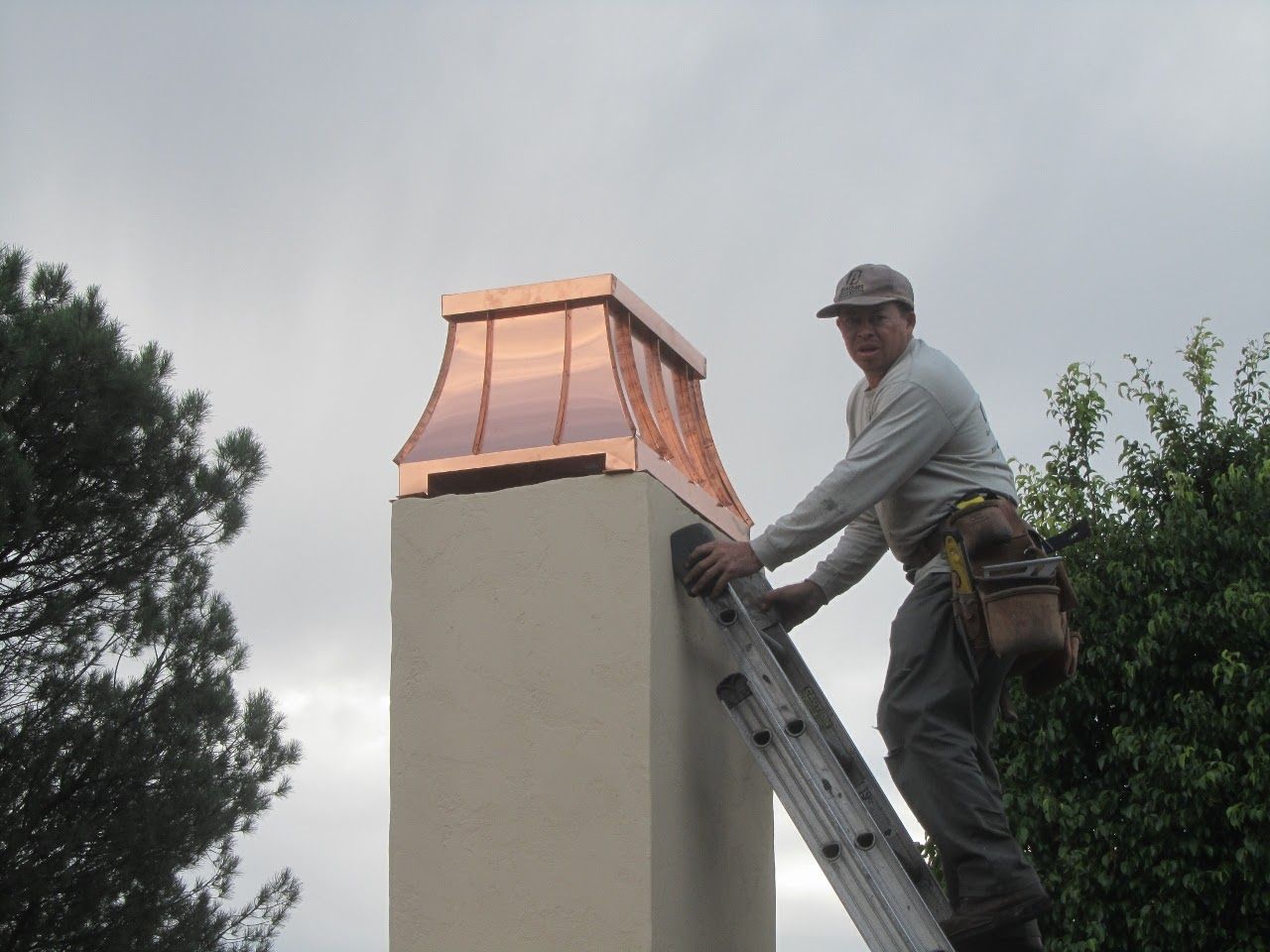 Man on a ladder installing copper chimney cap on a stucco chimney, outdoors. Cloudy sky, trees.