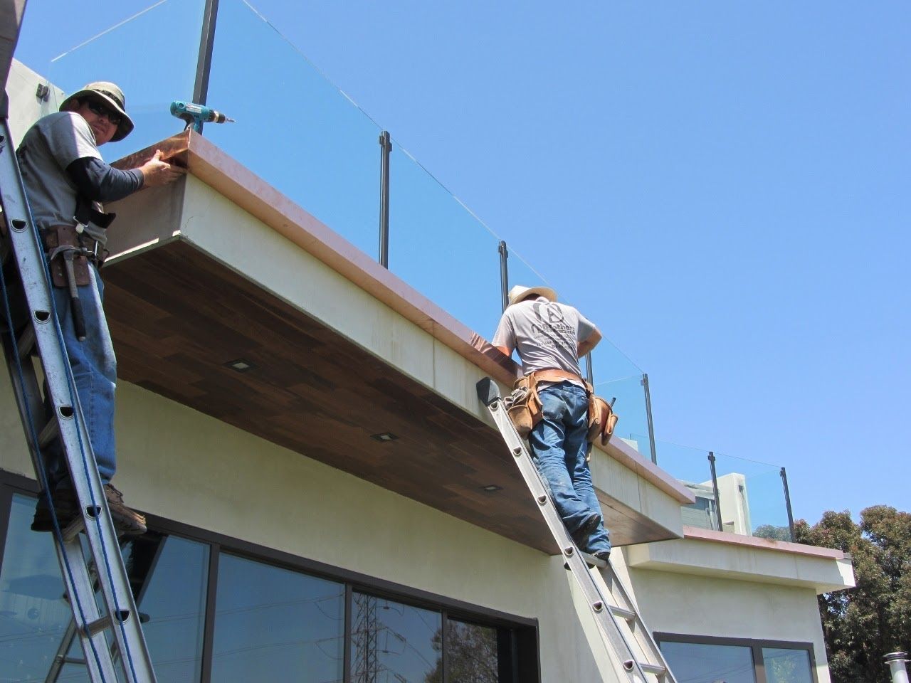 Two workers install a deck railing with glass panels. They stand on ladders, attaching the top rail. Sunny outdoor setting.