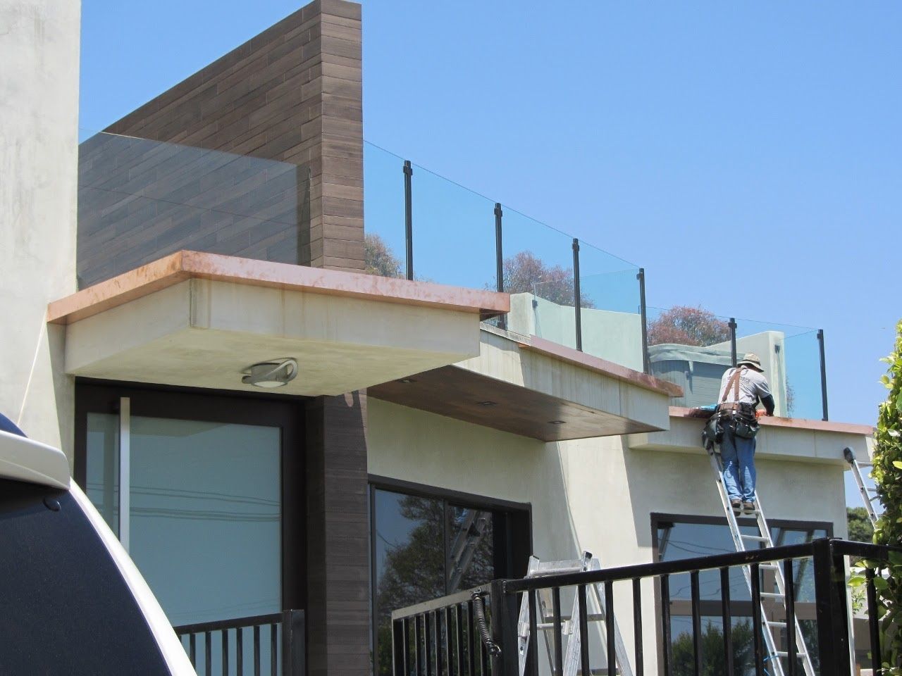 Person on a ladder working on a building's rooftop with glass railing. Blue sky, sunny day.