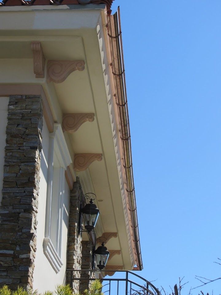 House exterior with stone siding, pink decorative brackets, and copper rain gutter against a blue sky.
