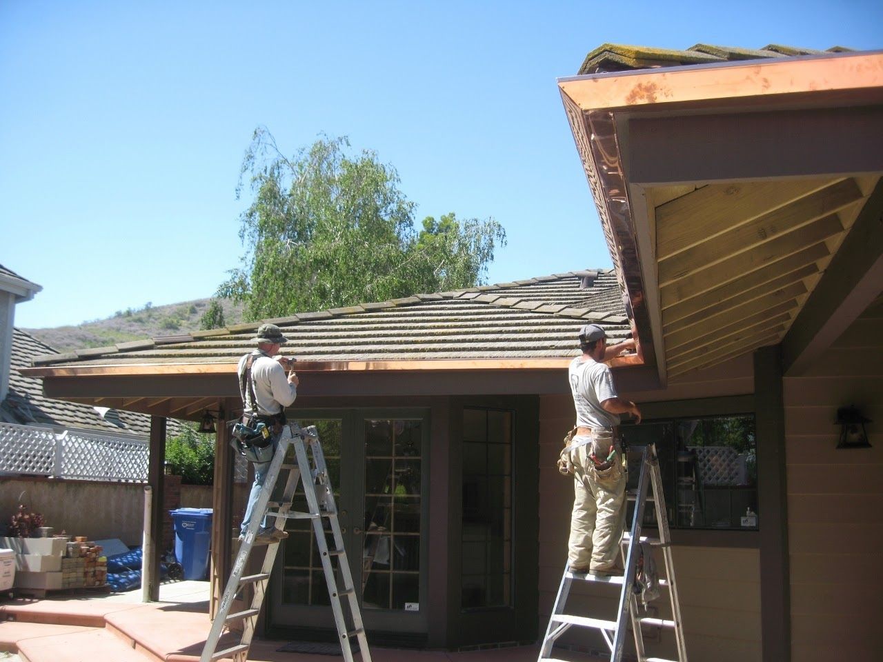 Two workers on ladders installing gutters on a house with a brown roof on a sunny day.