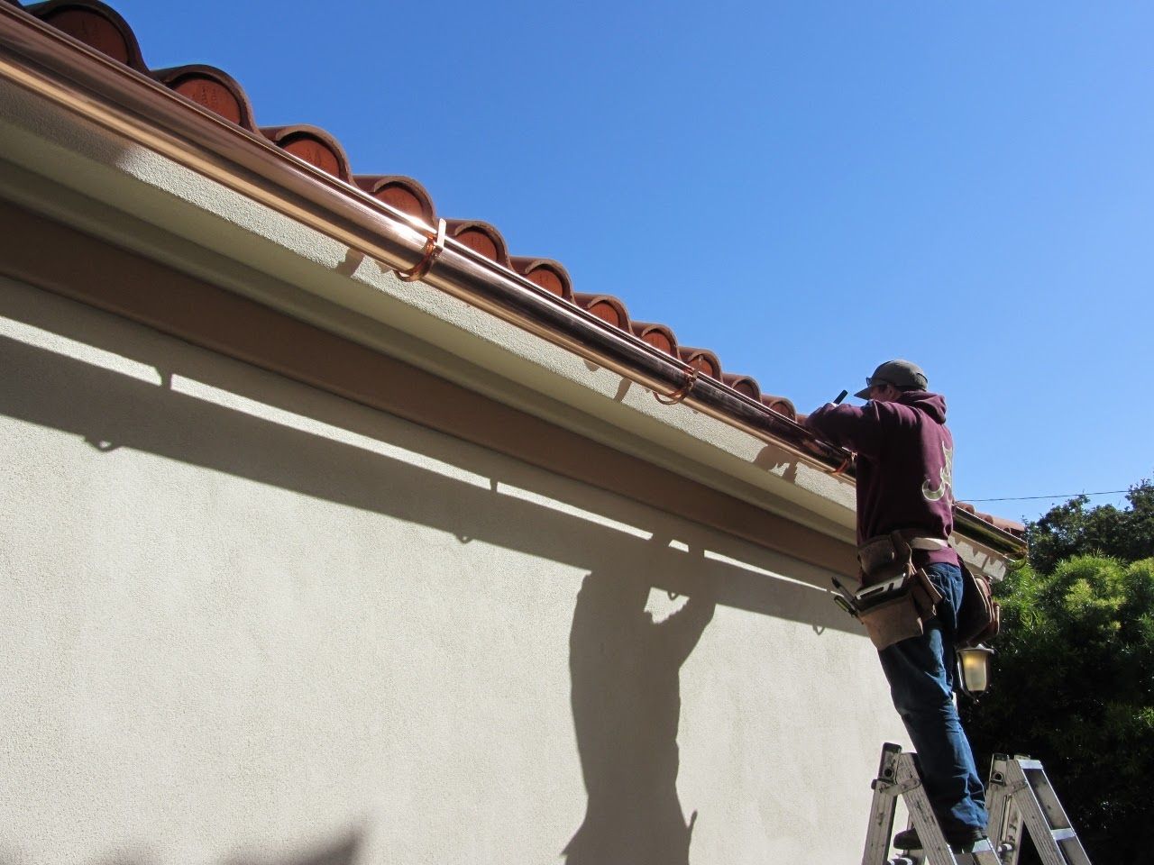 Person on ladder installing gutters on a house with terracotta roof tiles, against a blue sky.